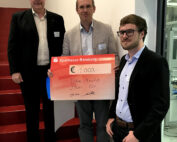 Drei Männer stehen auf einer Treppe und halten einen großen feierlichen Scheck über 1.000 Euro der Sparkasse Bamberg in den Händen, den Tobias Blaufuss für einen Wettbewerb erhalten hat. In Business-Kleidung feiern sie stolz den Erfolg an der Hochschule Coburg.