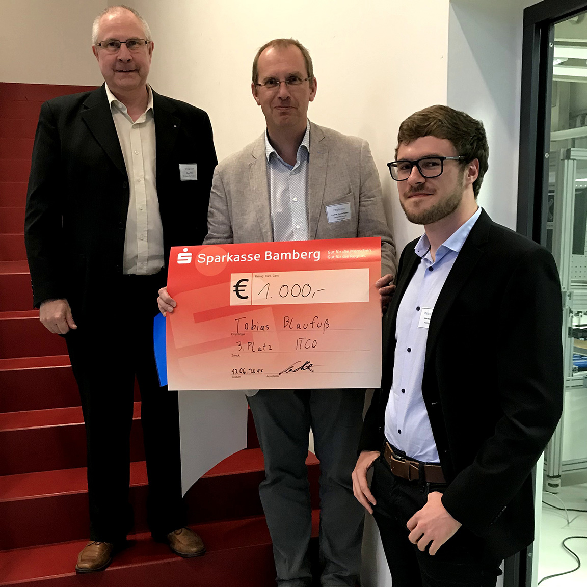 Drei Männer stehen auf einer Treppe und halten einen großen feierlichen Scheck über 1.000 Euro der Sparkasse Bamberg in den Händen, den Tobias Blaufuss für einen Wettbewerb erhalten hat. In Business-Kleidung feiern sie stolz den Erfolg an der Hochschule Coburg.