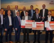 Eine Gruppe von Männern in formeller Kleidung, von denen einige die Hochschule Coburg repräsentieren, steht drinnen und hält große Schecks und Trophäen in den Händen. Im Hintergrund sind ein Feld und ein Sonnenuntergang zu sehen, was die festliche und professionelle Stimmung noch verstärkt. Einige Männer tragen Anzüge, während andere Blazer tragen.