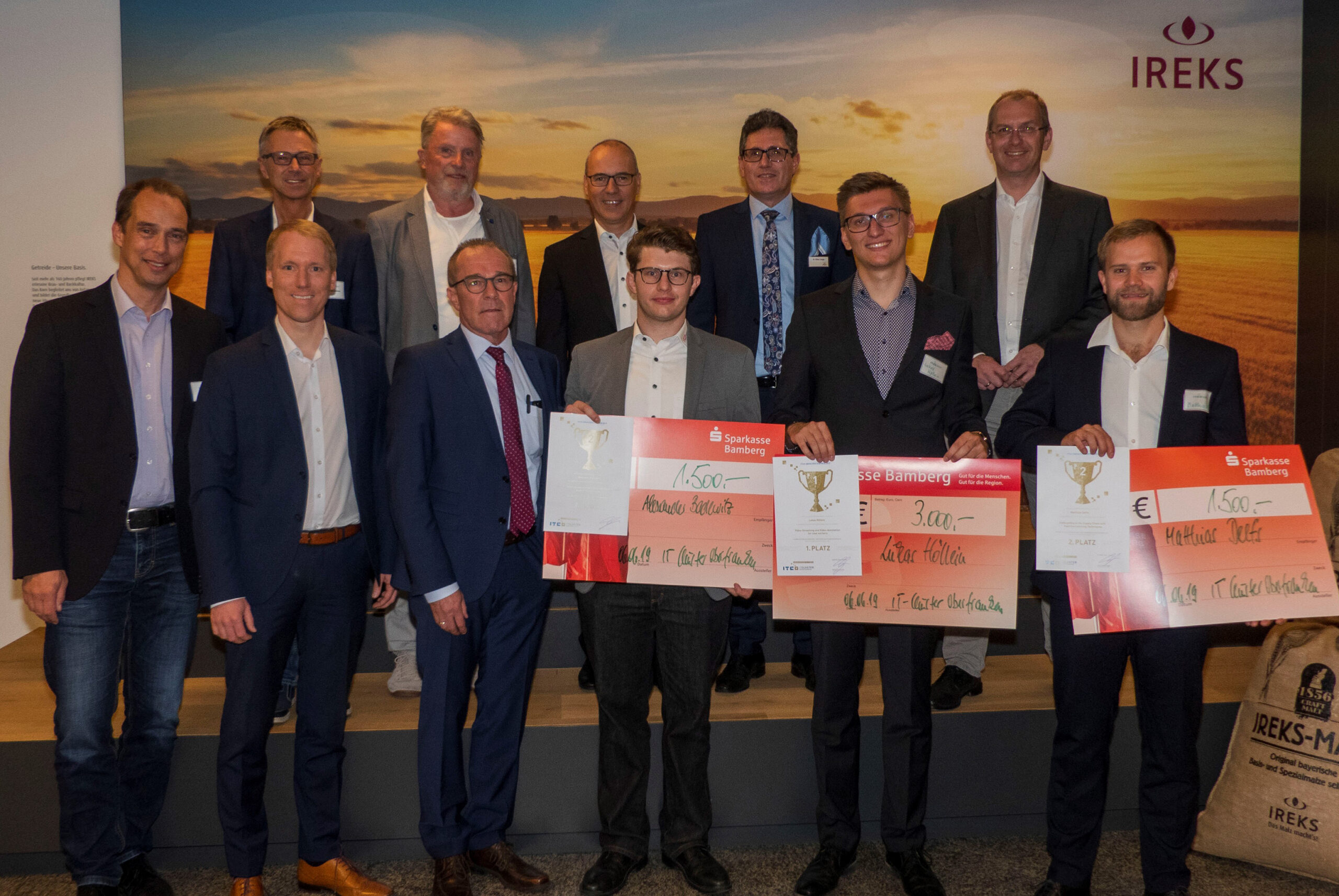 Eine Gruppe von Männern in formeller Kleidung, von denen einige die Hochschule Coburg repräsentieren, steht drinnen und hält große Schecks und Trophäen in den Händen. Im Hintergrund sind ein Feld und ein Sonnenuntergang zu sehen, was die festliche und professionelle Stimmung noch verstärkt. Einige Männer tragen Anzüge, während andere Blazer tragen.