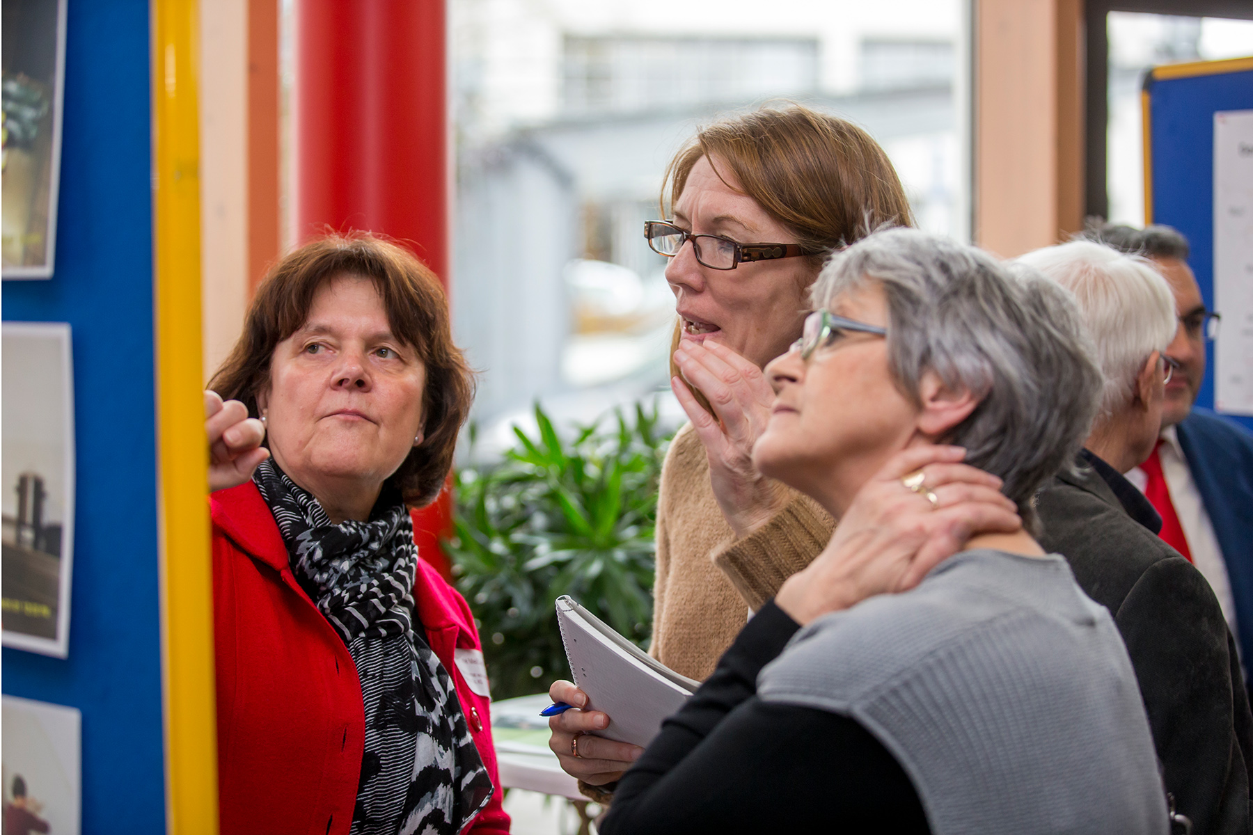 Drei Frauen unterhalten sich an einem schwarzen Brett der Hochschule Coburg. Eine zeigt auf das Brett, während die anderen aufmerksam zuhören. Sie befinden sich in einem Raum, mit einem unscharfen Hintergrund aus einer Pflanze und einem Fenster, was ihrer konzentrierten Diskussion eine gemütliche Atmosphäre verleiht.
