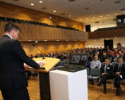 Ein Redner im Anzug spricht zu einem großen Publikum in einem modernen Konferenzsaal der Hochschule Coburg. Das Publikum hört aufmerksam zu, auf dem Podium liegen ein Laptop und Unterlagen. Der Raum ist mit Holzvertäfelung und Sitzreihen ausgestattet, was die akademische Atmosphäre verstärkt.