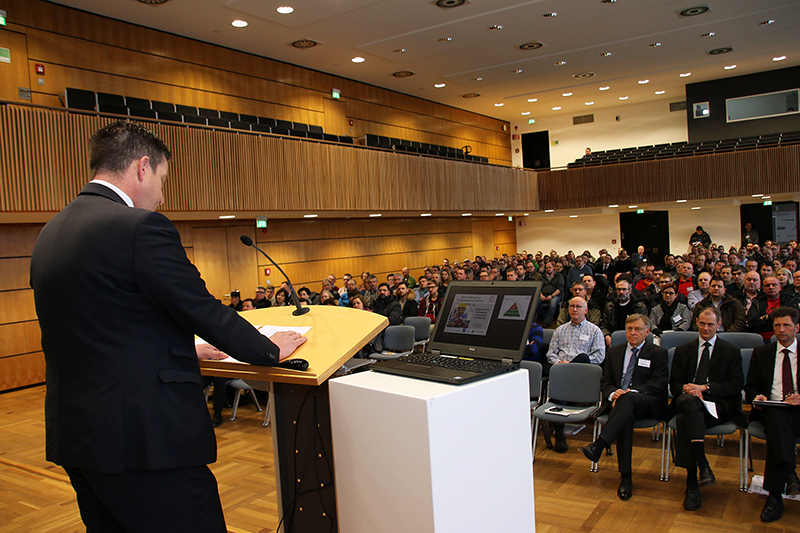 Ein Redner im Anzug spricht zu einem großen Publikum in einem modernen Konferenzsaal der Hochschule Coburg. Das Publikum hört aufmerksam zu, auf dem Podium liegen ein Laptop und Unterlagen. Der Raum ist mit Holzvertäfelung und Sitzreihen ausgestattet, was die akademische Atmosphäre verstärkt.