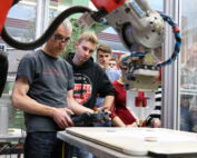 Eine Gruppe von Menschen an der Hochschule Coburg beobachtet einen Mann, der in einer Laborumgebung einen Roboterarm bedient. Der Mann konzentriert sich auf die Verwendung einer Handsteuerung und steuert die Maschine, während sie auf einer flachen Oberfläche arbeitet, während andere aufmerksam zuschauen.