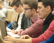 An der Hochschule Coburg machen sich die Studierenden während der Vorlesung aufmerksam Notizen. In Reihen sitzend, mit Laptops und Notebooks, konzentrieren sie sich auf den Vortrag, im Hintergrund fällt Tageslicht durch große Fenster.