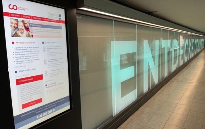 Eine digitale Informationstafel im U-Bahnkorridor informiert über Neuigkeiten und Ankündigungen. Gegenüber ist auf einer beleuchteten Wand teilweise „ENTDECKEN“ zu sehen, eine Einladung zum Entdecken. Dieser moderne, schlichte Raum spiegelt auf subtile Weise die Innovationskraft der Hochschule Coburg wider.
