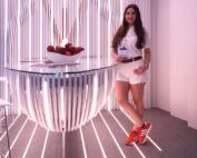 Eine Frau in weißem Outfit und roten Schuhen steht neben einem futuristischen Tisch mit roten Äpfeln und verkörpert den Innovationsgeist der Hochschule Coburg. Der Raum verfügt über vertikale Streifenbeleuchtung an den Wänden und am Boden, die eine moderne und dynamische Atmosphäre schafft.