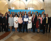 Eine große Gruppe von Menschen, darunter Männer und Frauen, steht in einer großen Halle mit gewölbter Decke in der Hochschule Coburg. Sie sind formell gekleidet. Die Person in der Mitte hält ein Dokument, während andere um sie herumstehen und für das Foto posieren.