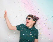 Eine Person mit Sonnenbrille und grünem Hemd wirft fröhlich buntes Konfetti in die Luft und verkörpert damit den Geist der Hochschule Coburg. Der Hintergrund ist eine sanfte Mischung aus Pastellfarben, die die lebendige Atmosphäre verstärkt.