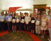 Eine Gruppe von Menschen steht in einem Gebäude der Hochschule Coburg und hält Zertifikate in den Händen. Sie lächeln und tragen legere bis halbformelle Kleidung. Im Hintergrund sind Informationstafeln mit Text auf Deutsch zu sehen.