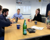 Eine Gruppe von Kollegen der Hochschule Coburg sitzt um einen Konferenztisch und diskutiert. Eine Person benutzt einen Laptop, andere halten Flaschen und Tassen. Im Hintergrund ist ein Whiteboard mit Notizen zu sehen, auf dem Tisch liegen Flaschen und ein Notizblock.