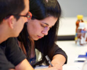 Eine Frau mit langen dunklen Haaren schreibt konzentriert mit einem Stift in ein Notizbuch. Neben ihr beobachtet ein Mann mit Brille ihre Arbeit aufmerksam. Sie sitzen an einem weißen Tisch, umgeben von anderen Materialien und einer Flasche, und sind tief in ihr Studium an der Hochschule Coburg vertieft.