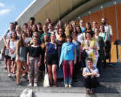 Eine große Gruppe leger gekleideter und lächelnder Menschen versammelt sich auf den sonnenbeschienenen Stufen vor einem modernen Gebäude. Einige sitzen, andere stehen und halten einen Moment an der Hochschule Coburg fest.