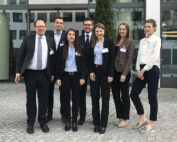 Eine Gruppe von sieben professionell gekleideten Personen steht vor einem Gebäude der Hochschule Coburg und lächelt in die Kamera. Sie tragen Business-Kleidung, darunter Anzüge und Blazer. Im Hintergrund sind Fenster und eine gepflasterte Fläche zu sehen.