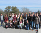 Eine bunt gemischte Gruppe von 27 Personen, die mit der Hochschule Coburg verbunden sind, posiert an einem sonnigen Tag im Freien. Sie stehen und hocken auf einer gepflasterten Fläche mit Bäumen und Hügeln im Hintergrund, lächeln und sind leger gekleidet.