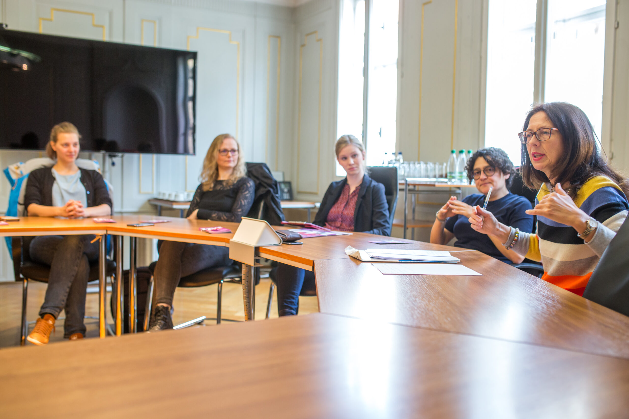 Eine Gruppe von fünf Personen sitzt um einen hölzernen Konferenztisch in einem hellen Raum der Hochschule Coburg. Vier Frauen hören aufmerksam zu, während eine Frau spricht und mit den Händen gestikuliert. Im Hintergrund sind Büromaterialien und Wasserflaschen zu sehen.