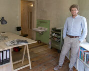 Ein Mann in einem blauen Hemd steht in einem kleinen, minimalistischen Büro mit Holzboden an der Hochschule Coburg. Der Raum verfügt über einen Schreibtisch mit einem aufgeschlagenen Buch, eine Lampe und Regale voller Bücher. Im Hintergrund vervollständigen ein Waschbecken und eine Tür die ruhige akademische Atmosphäre.