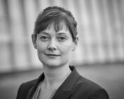 Schwarz-Weiß-Porträt einer Person mit kurzen Haaren und Pony, die einen Blazer der Hochschule Coburg trägt und vor einem unscharfen Hintergrund steht.