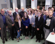 Eine Gruppe von Menschen unterschiedlichen Alters steht in einem gut beleuchteten Raum der Hochschule Coburg. Einige sind leger gekleidet, andere tragen Business-Kleidung. Sie scheinen sich in einem akademischen oder beruflichen Umfeld zu befinden, im Hintergrund sind Tische und Geräte zu sehen.