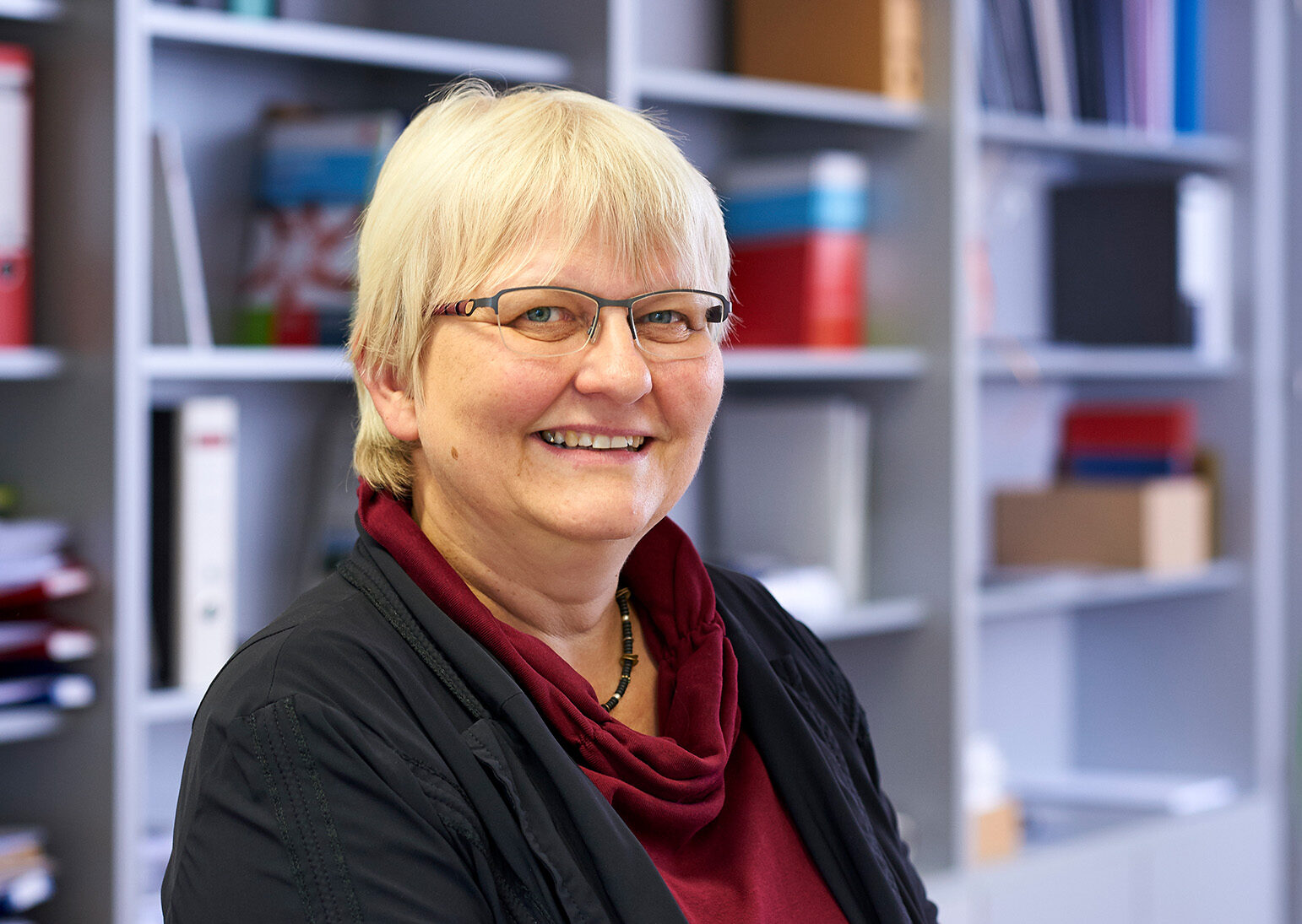 Eine Person mit kurzen blonden Haaren und Brille steht lächelnd vor einem Regal voller Bücher und Ordner in der Hochschule Coburg. Sie trägt eine schwarze Jacke und ein kastanienbraunes Oberteil.