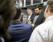 Eine Gruppe von Menschen in einem Raum, wobei eine Person einen Metallgegenstand hält und möglicherweise dessen Funktion erklärt. Sie sind in Business-Kleidung gekleidet und befinden sich in einem professionellen Umfeld, wahrscheinlich einem Büro oder einer Werkstatt an der Hochschule Coburg.