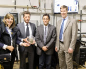 Vier Personen stehen in einem Raum der Hochschule Coburg, im Hintergrund sind Industrieanlagen und Rohre zu sehen. Eine Person hält eine transparente Auszeichnung in der Hand. Sie sind alle in formeller Kleidung gekleidet und lächeln in die Kamera.