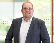 Ein Mann mit Brille steht in einem Gebäude der Hochschule Coburg. Er trägt einen dunklen Blazer über einem weißen Hemd. Vor dem hellen Hintergrund unscharfer Fenster blickt er mit neutralem Gesichtsausdruck in die Kamera.