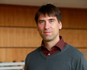 Eine Person mit kurzen braunen Haaren, die einen grauen Pullover über einem kastanienbraunen Hemd trägt, steht vor einer holzgetäfelten Wand in der Hochschule Coburg und lächelt leicht in die Kamera.