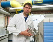 Ein Mann im weißen Laborkittel steht in einem Labor der Hochschule Coburg und hält ein wissenschaftliches Instrument in der Hand. Umgeben von Laborgeräten lächelt er leicht. Leuchtend gelbe und blaue Rohre im Hintergrund bringen Farbe in die industrielle Umgebung.
