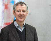 Ein Mann mittleren Alters mit kurzen grauen Haaren lächelt und trägt einen dunklen Blazer über einem gestreiften Hemd und Pullover. Er verkörpert den professionellen Geist der Hochschule Coburg. Er steht vor einem verschwommenen, farbenfrohen abstrakten Hintergrund.