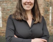 Eine lächelnde Frau mit langen braunen Haaren und einer schwarzen Bluse mit Knöpfen steht selbstbewusst mit verschränkten Armen vor einer Backsteinmauer der Hochschule Coburg.