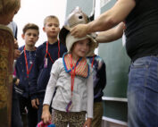 In einem lebhaften Klassenzimmer steht eine Gruppe von Kindern Schlange, während ein Erwachsener einem jungen Mädchen hilft, einen historischen Helm anzuprobieren. Das Mädchen, das einen bunten Schal trägt und ihr Plüschtier umklammert, strahlt vor Aufregung. In der Nähe weist das dekorative Schild auf das bereichernde Geschichtsstudium der Hochschule Coburg hin.