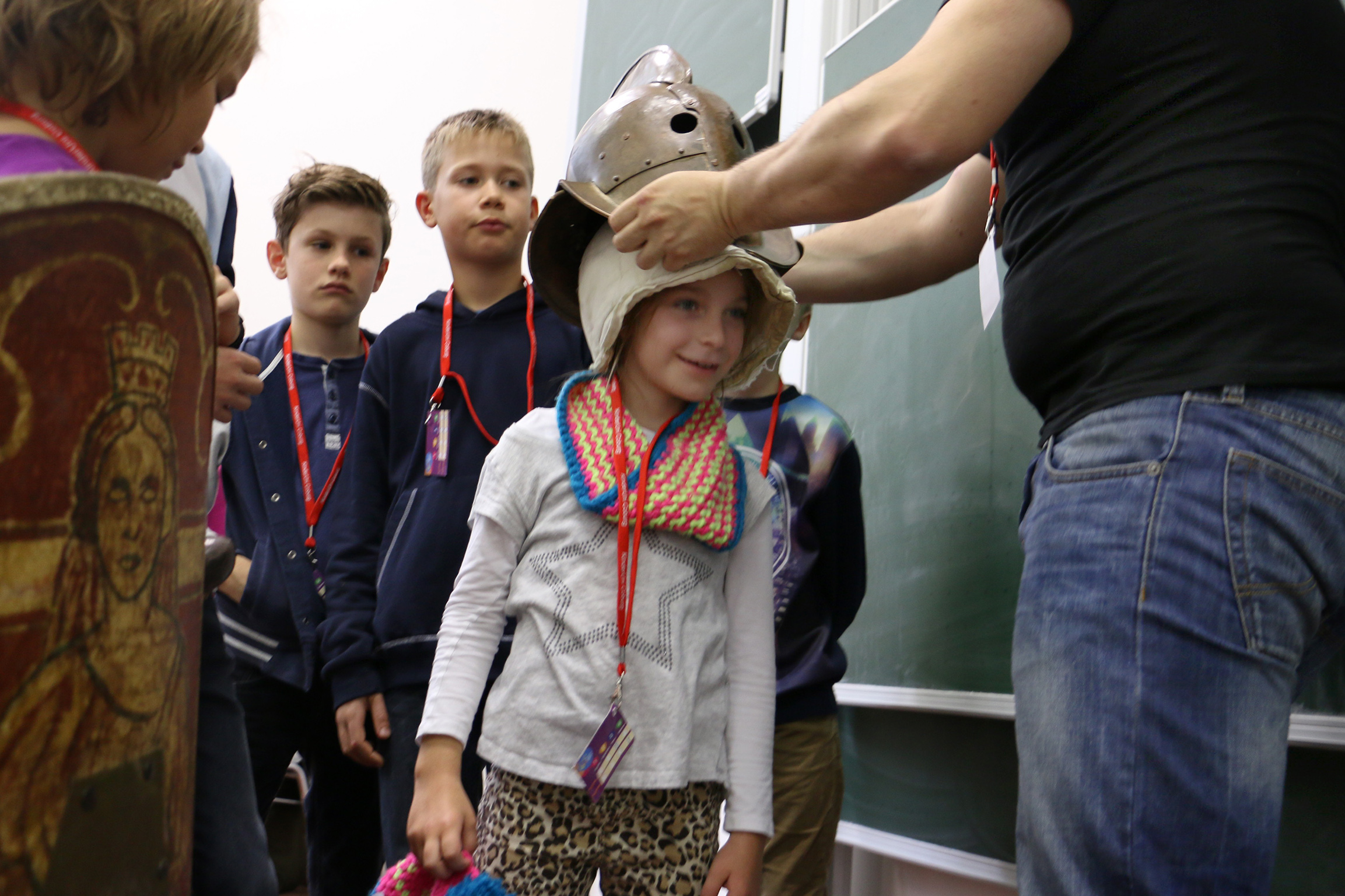 In einem lebhaften Klassenzimmer steht eine Gruppe von Kindern Schlange, während ein Erwachsener einem jungen Mädchen hilft, einen historischen Helm anzuprobieren. Das Mädchen, das einen bunten Schal trägt und ihr Plüschtier umklammert, strahlt vor Aufregung. In der Nähe weist das dekorative Schild auf das bereichernde Geschichtsstudium der Hochschule Coburg hin.