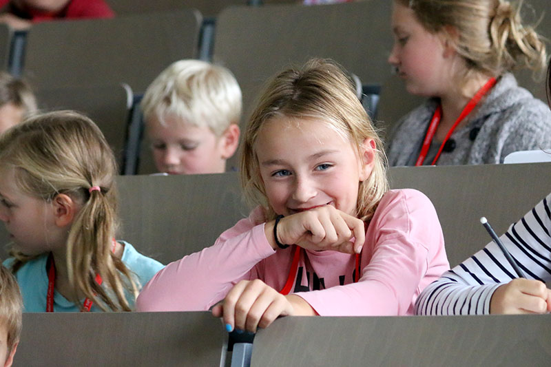 Kinder sitzen in einem Hörsaal der Hochschule Coburg und sind mit einer Aktivität beschäftigt. Ein Mädchen in einem rosa Hemd lächelt und legt seinen Arm auf den Tisch, umgeben von anderen Kindern mit roten Schlüsselbändern.