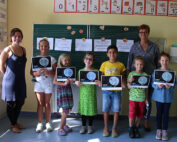 Eine Gruppe von sieben Kindern steht lächelnd in einem Klassenzimmer der Hochschule Coburg. Jedes Kind hält ein Zertifikat in der Hand. Zwei Erwachsene stehen neben ihnen und strahlen vor Stolz. Hinter ihnen ist eine Tafel mit Zeichnungen und Texten. Der Raum ist gut beleuchtet und mit pädagogischen Plakaten dekoriert.