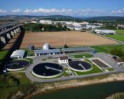 Luftaufnahme der Kläranlage in der Nähe der Hochschule Coburg mit runden Tanks und angrenzenden Gebäuden. Eine Autobahnüberführung verläuft daneben, mit offenen Feldern und entfernten Hügeln im Hintergrund unter einem teilweise bewölkten Himmel.