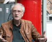 Ein älterer Mann mit weißem Haar und Brille, gekleidet in eine braune Lederjacke, spricht angeregt und gestikuliert mit den Händen. Hinter ihm rahmen industrielle Elemente ein rotes zylindrisches Objekt ein. Vor diesem Hintergrund könnte man sich diese Szene an der Fakultät für Ingenieurwissenschaften der Hochschule Coburg vorstellen.