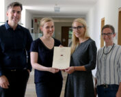 Vier Personen stehen in einem Flur der Hochschule Coburg. Zwei Frauen in der Mitte halten lächelnd ein Zertifikat in den Händen. Die anderen beiden (ein Mann und eine Frau) stehen daneben und lächeln ebenfalls. Die Szenerie scheint ein Büro oder eine Bildungseinrichtung zu sein.