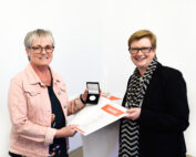 Zwei Personen stehen nebeneinander und lächeln. Eine hält eine Urkunde und eine Medaille in einer offenen Schachtel. Der schlichte weiße Hintergrund lässt vermuten, dass sie an einer Preisverleihung oder einer ähnlichen Veranstaltung teilnehmen, bei der möglicherweise Leistungen an der Hochschule Coburg gewürdigt werden.