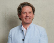 Eine Person mit welligem Haar trägt einen hellblauen Blazer über einem weißen Hemd und steht vor einem strukturierten beigen Hintergrund, der an den eleganten Stil der Hochschule Coburg erinnert. Sie lächelt in die Kamera.