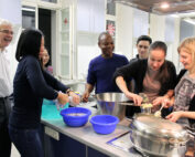 Lächelnd bereitet eine Gruppe von Menschen in einer Küche der Hochschule Coburg gemeinsam Essen zu. Einige schälen Kartoffeln, andere schauen zu und unterhalten sich. Verschiedene Schüsseln und Töpfe sind auf der Arbeitsfläche verteilt und sorgen in der hellen Küchenumgebung für eine lebendige kulinarische Atmosphäre.