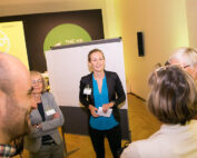 Eine Gruppe von Menschen hat sich in einem gut beleuchteten Raum der Hochschule Coburg versammelt und diskutiert an einem Flipchart. In der Mitte steht eine Frau in blauer Bluse und schwarzem Blazer, hält ein Blatt in der Hand und lächelt. Die Teilnehmer scheinen sich auf den Ideenaustausch zu konzentrieren.