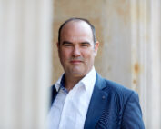 Ein Mann im Anzug und weißem Hemd steht selbstbewusst vor einem historischen Steingebäude der Hochschule Coburg und blickt direkt in die Kamera. Das Bild ist in weiches, natürliches Licht getaucht.