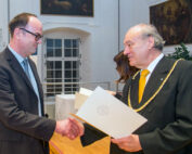 Zwei Männer in formeller Kleidung geben sich bei einer Preisverleihung die Hand. Einer trägt eine Dienstkette und hält eine Urkunde in der Hand. Sie stehen in einem Raum mit Holzfußboden und Gemälden, möglicherweise als Zeichen einer Veranstaltung an der Hochschule Coburg.