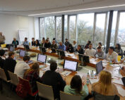 In einem Konferenzraum der Hochschule Coburg sitzt eine Gruppe von Menschen mit Papieren und Monitoren an rechteckigen Tischen. Große Fenster geben den Blick auf die Bäume draußen frei. Vorne steht ein Redner neben einem Whiteboard und spricht zu den Teilnehmern.