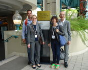 Vier Menschen stehen in der hell erleuchteten Lobby des Victoria Conference Centre zusammen. Sie lächeln und tragen Namensschilder mit der Aufschrift „Hochschule Coburg“. Die moderne Einrichtung und die Pflanzen schaffen eine einladende Atmosphäre, als sie sich unter einem Willkommensschild versammeln.