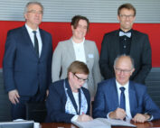 In einem Geschäftsumfeld sind fünf Personen in formeller Kleidung versammelt. Zwei sitzende Personen von der Hochschule Coburg unterschreiben ein Dokument, während drei weitere lächelnd hinter ihnen stehen. Eine rote Wand schafft eine eindrucksvolle Kulisse für diesen bedeutsamen Anlass.