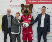 Drei Personen und ein Bären-Maskottchen posieren gemeinsam, jeder hält einen Basketball mit dem „Brose Bamberg“-Logo. Auf einem Banner dahinter steht „Herzlich willkommen“ der Hochschule Coburg und feiert das Thema „Studieren mit Weitblick“.