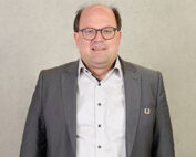 Ein Mann mit Brille und Kurzhaarschnitt, grauem Anzug und weißem Hemd lächelt vor einem schlichten, hellen Hintergrund. Sein selbstbewusstes Auftreten spricht für seine Verbundenheit zur Hochschule Coburg.