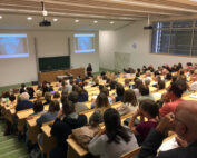 Im voll besetzten Hörsaal der Hochschule Coburg hält ein Redner einen Vortrag vor einem aufmerksamen Publikum. Dank der abgestuften Holzsitze und zwei Bildschirmen, auf denen das gleiche Bild gezeigt wird, können sich die Teilnehmer in einem hellen, modernen Ambiente auf die Präsentation konzentrieren.